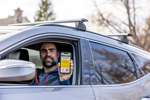 Vaccinated man using digital vaccine passport app in mobile phone at customs for travel during Covid-19 pandemic. He is showing his immunization certificate and passport to the customs official. He is sitting in his car. French : Certificat International de Vaccination