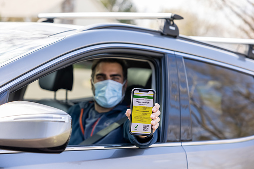 Vaccinated man using digital vaccine passport app in mobile phone at customs for travel during Covid-19 pandemic. He is showing his immunization certificate and passport to the customs official. He is sitting in his car. He is wearing a protective face mask.