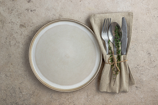 Plate and cutlery with linen napkin on beige stone backgroud. Rustic table setting in natural warm colors. Top view. Flat lay