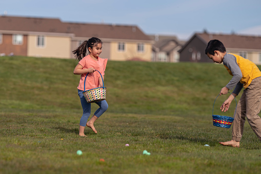 Mixed race elementary age brother and sister race barefoot through the grass on a warm, sunny day to pick up eggs and candy during an Easter egg hunt. The barefoot kids are celebrating Easter outdoors in their neighborhood. Suburban homes and townhouses are visible in the background.