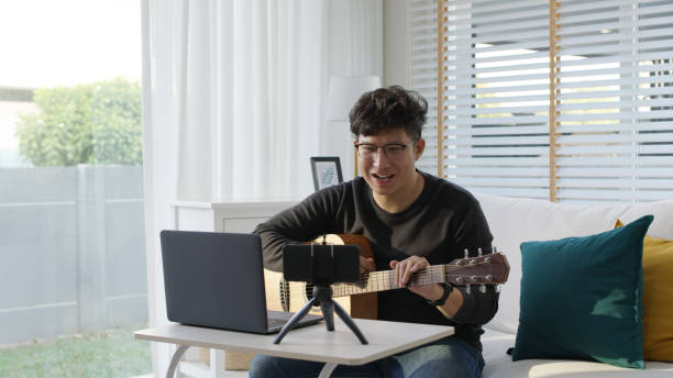 asiatische gitarrist livestream-video oder virtuelle video-tutorial zu hause, künstler musiker blogger oder vlogger sitzen auf dem sofa im wohnzimmer erklären, gitarre zu spielen, online-lernen während quarantäne-konzept - men artist guitarist guitar stock-fotos und bilder