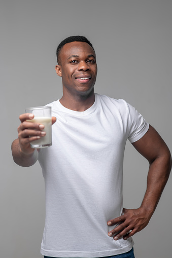Proper nutrition. Cheerful young good looking dark skinned man holding out glass of milk forward in great mood