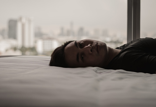 Asian man feels sad alone on bed with city background. Stay home, depression and loneliness concept.