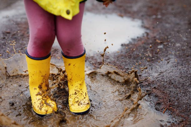 primo primo tempo di una bambina che indossa stivali da pioggia gialli e cammina durante il nevischio nella giornata nuvolosa piovosa. bambino carino in abiti colorati che salta nella pozzanghera, spruzza con acqua, attività all'aperto - dirt jumping foto e immagini stock