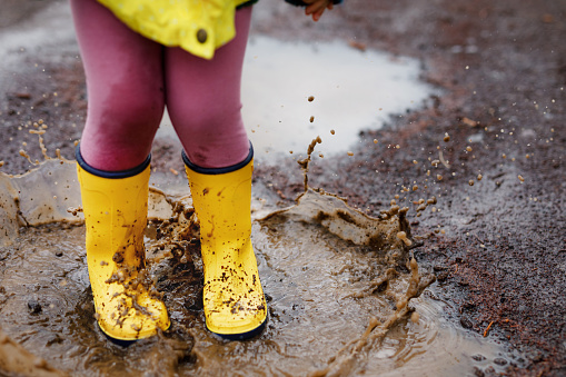 Close-up of little toddler girl wearing yellow rain boots and walking during sleet on rainy cloudy day. Cute child in colorful clothes jumping into puddle, splashing with water, outdoor activity.