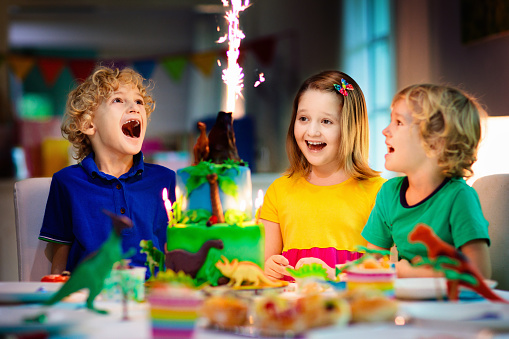 Kids birthday party. Dinosaur theme cake. Little girl blowing candles and opening gifts. Children event. Decoration for dinosaurs themed celebration.