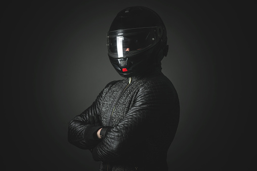 Serious motorbiker in a helmet with a crossed arms is standing on the dark background.