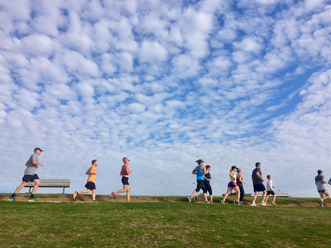 Horizontal landscape of people, young and old, jogging along a coastal beach promenade in Victoria on a sunny Summer day under a blue sky with white clouds