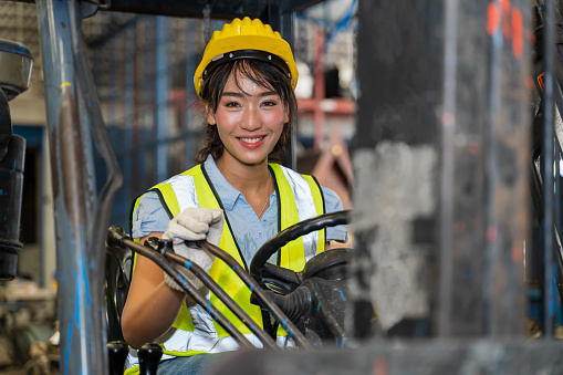 A female engineer is controlling a forklift in a spare parts factory. A smiling woman is deliberately practicing machine control in a factory in Industrial Engineering worker concept