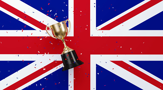 Star shaped gold confetti falling onto a gold cup sitting over British flag background. Horizontal composition with copy space. Front view. Championship concept.