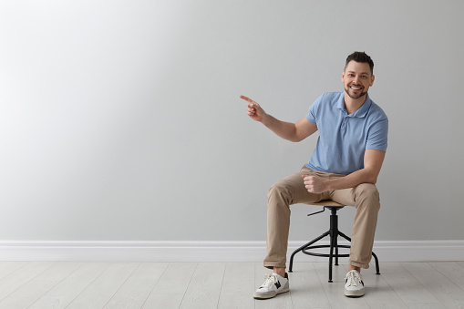 Handsome man sitting in office chair near grey wall indoors, space for text