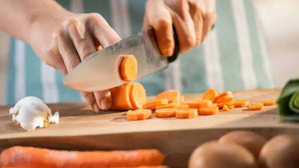 Close-up of female hands cutting carrot with knife on chopping board in kitchen.
