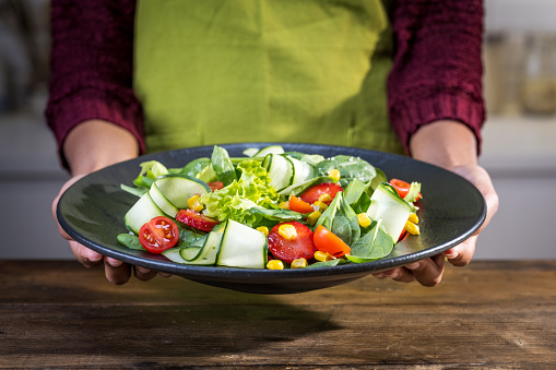 Close-up of woman's hands holding plate of mixed vegetable salad in kitchen.