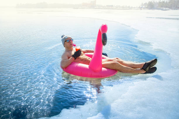 A man swimming in an ice hole in winter in Finland, A man swimming in an ice hole in winter in Finland, floating on a pink inflatable flamingo with cocktail in hand. Vacation options, dreaming of summer. bizarre stock pictures, royalty-free photos & images