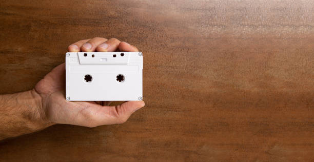 There is a white audio magnetic cassette in the man's hand. Below is a wooden brown background. There is a white audio magnetic cassette in the man's hand. Below is a wooden brown background. Copy Space. reel to reel tape stock pictures, royalty-free photos & images