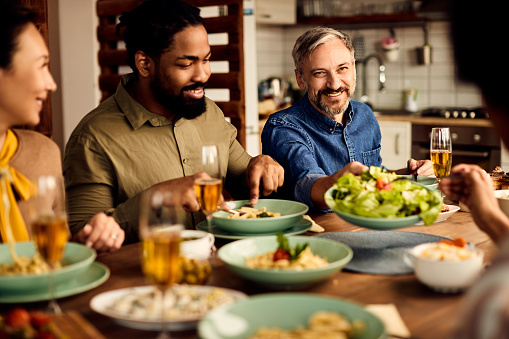Happy man having lunch with friends and offering salad to one of them at dining table.