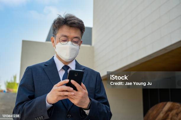 Middleaged Asian Businessman In The City Wearing A Mask And Using A Smartphone Stock Photo - Download Image Now