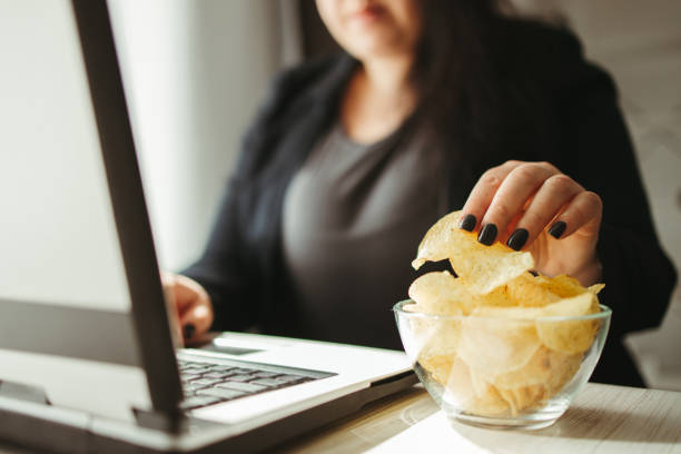 Woman eating junk food, snacking with chips Compulsive overeating, mindless snacking, junk food, unhealthy meals. Woman eating chips from bowl at her workplace unhealthy eating stock pictures, royalty-free photos & images