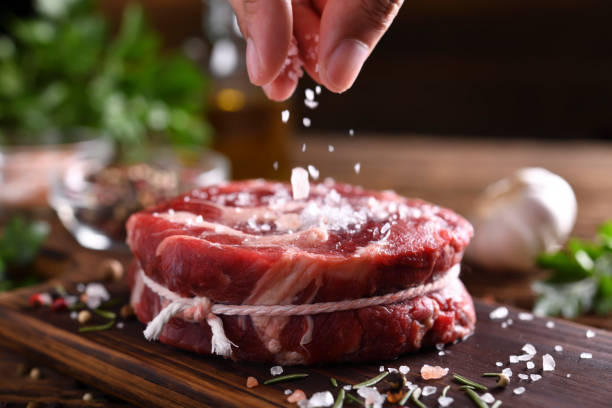 handbestreuen von salz auf frischem rohem rindfleischfleisch auf einem schneidebrett - streuen fotos stock-fotos und bilder