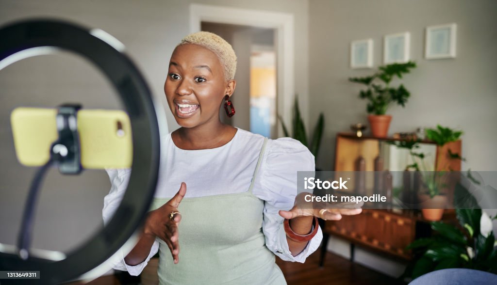 Smiling young African female influencer doing a vlog post at home Smiling young African female influencer standing in her living room at home and talking during a vlog post using a smart phone Influencer Stock Photo