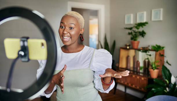 Smiling young African female influencer doing a vlog post at home Smiling young African female influencer standing in her living room at home and talking during a vlog post using a smart phone social media marketing stock pictures, royalty-free photos & images