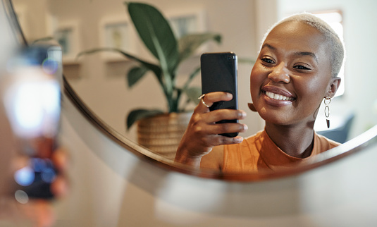 Young African woman with short blond hair smiling and taking selfies with her smart phone in front of a mirror at home