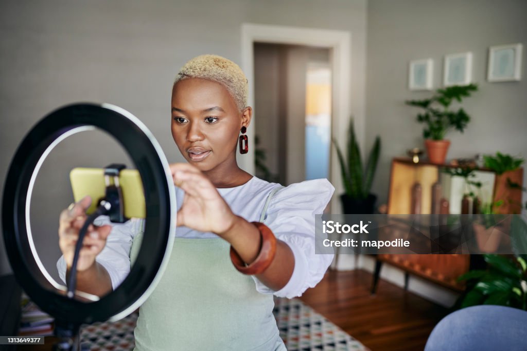 Young African influencer adjusting her smart phone before a vlog post Young African female influencer adjusting a smart phone and ring light before doing an online vlog post at home Influencer Stock Photo