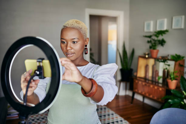 Young African influencer adjusting her smart phone before a vlog post Young African female influencer adjusting a smart phone and ring light before doing an online vlog post at home tutorial stock pictures, royalty-free photos & images