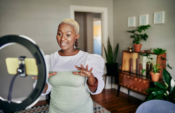 Smiling African influencer doing a vlog post in her living room Smiling young African female influencer talking during a vlog post using a smart phone while standing in her living room at home plus size photos stock pictures, royalty-free photos & images