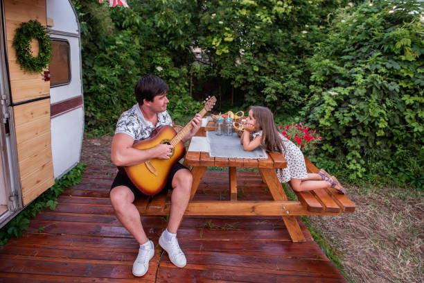 młody brunetka ojciec gra piosenki gitara dla małej pięknej córki. mężczyzna i dziewczyna siedzą przy drewnianym stole na podwórku, ciesząc się muzyką. ciężarówka z przyczepą z wakacji rodzinnych poza miastem - class a motorhome zdjęcia i obrazy z banku zdjęć