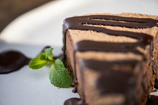 Close-up of slice of chocolate cake with brown fudge sauce and mint leaf served on plate.