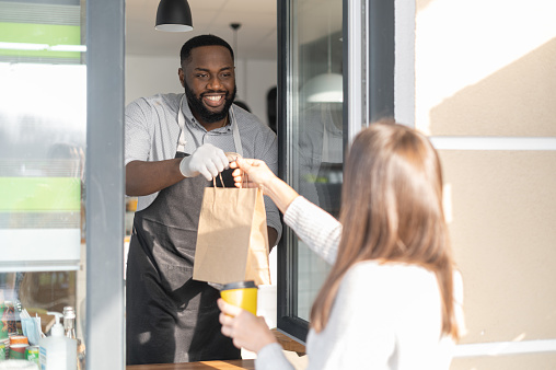 A friendly and smiling African-American waiter is serving female customer, a multiracial cafe manager holds out a take-away order through take-and-go window