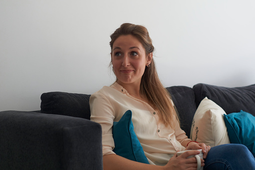 Beautiful young woman sitting on an armchair, looking carefree and having a cup of coffee