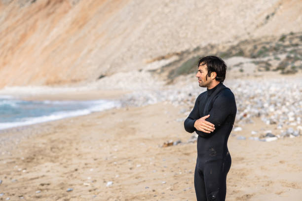 istock Side view medium shot wet man with diving suit and folded arms standing on the beach and looking at distance. 1313666767