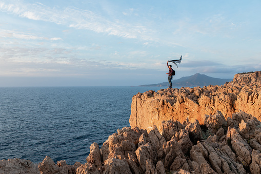 Young man standing on a high jagged cliff waving a scarf. Looking the sunset on the sea.