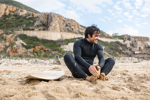 Young surfer man wearing diving suit sitting on the sand next to his surfboard smiling and resting after surfing.