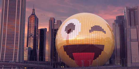 A huge spherical building covered with coloured glass to the form a yellow emoji face which is blinking and smiling with tongue out. The built structure is in the middle of tall downtown skyscrapers at sunset or sunrise. There is solitary car on the overpass and some birds in flight.