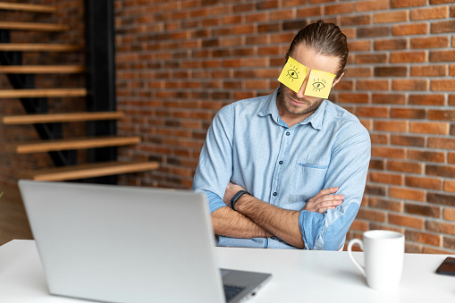 A young tired bearded office worker in a casual shirt sitting at the desk at his workplace, put stickers on the eyes to hide that he is napping or sleeping during work time, folded crossed his arms