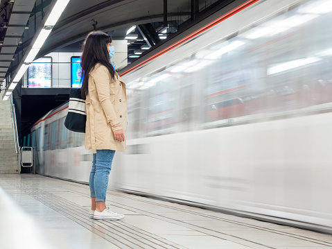 Woman standing on a subway platform with a metro train moving and arriving at the underground station. A young girl waiting for public transport during the covid pandemic.