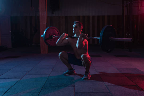 istock Working out by lifting weights in a cross-training gym. Muscular powerful man with heavy barbell doing squat exercise in red blue neon light 1313692870