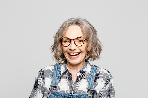 Excited elderly lady wearing dungarees and checkered shirt standing against grey background, laughing at camera. Studio shot of female designer.