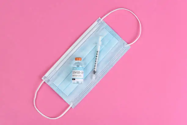 Equipment to administer a Covid19 Vaccination. Flat lay with surgical mask, syringe and vaccine vial, on pink background.