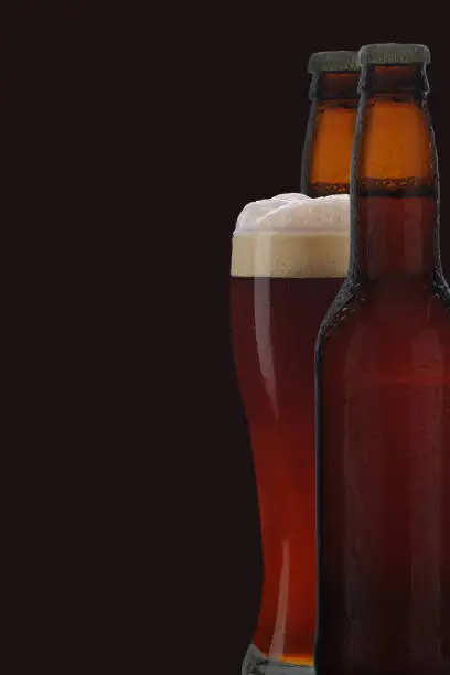 A glass of dark beer between two brown beer bottles against black background.