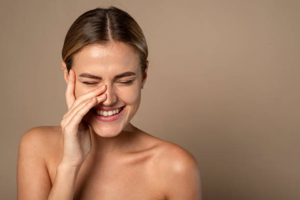 cuidado de la piel. mujer con cara de belleza tocando piel facial saludable. hermosa modelo femenina caucásica sonriente con maquillaje natural tocando la piel hidratada brillante sobre fondo beige - spa treatment fotos fotografías e imágenes de stock