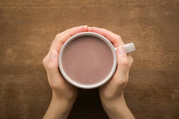 Photo of Young adult woman hands holding cup of cocoa on dark brown wooden table background. Closeup. Point of view shot. Top down view.