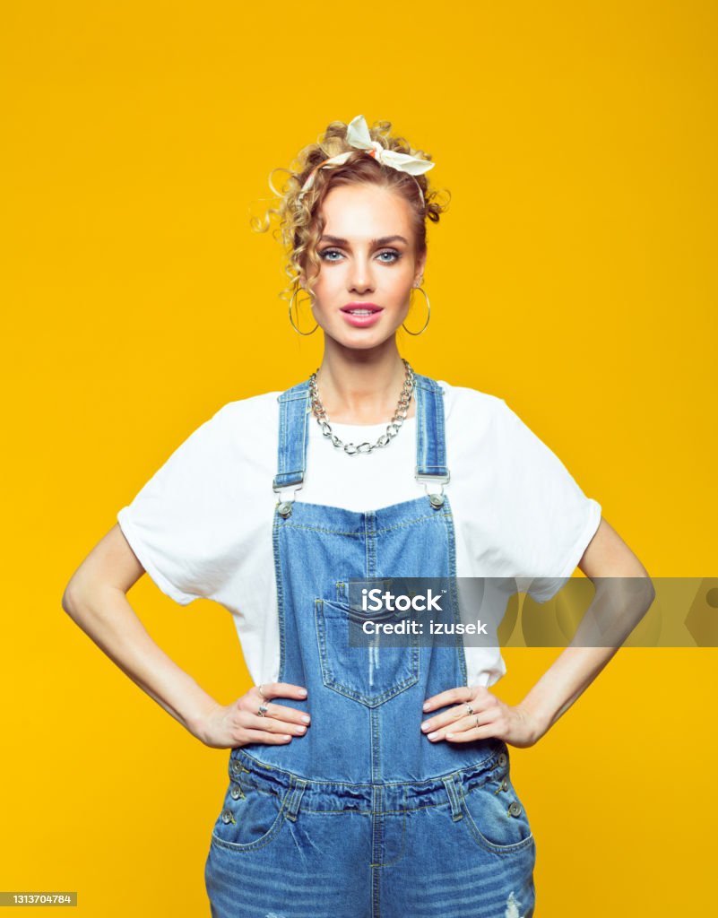 Portrait of confident young woman in 80's style outfit Portrait of young woman wearing white t-shirt, denim dungarees and bandana looking at camera with hands on hips. Studio shot on yellow background. 1980-1989 Stock Photo