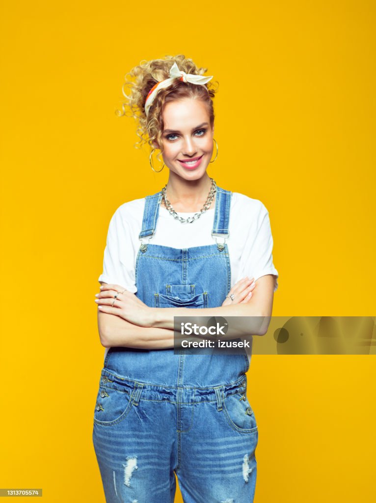 Portrait of confident young woman in coveralls on yellow background Portrait of young woman wearing white t-shirt, denim dungarees and bandana looking at camera with arms crossed. Studio shot on yellow background. Authority Stock Photo