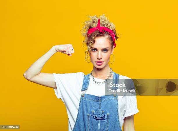 Powerful Young Woman In Dungarees Raising Fist Portrait On Yellow Background Stock Photo - Download Image Now