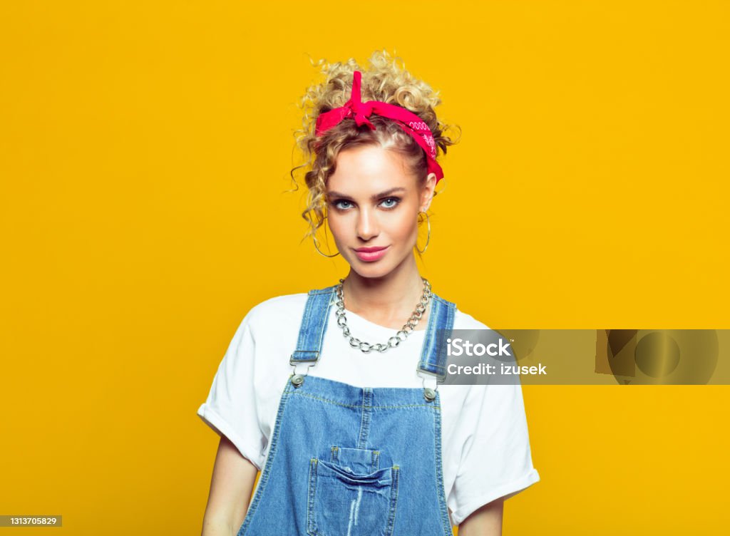 Young woman in 80's style outfit, portrait on yellow background rtrait of happy young woman wearing white t-shirt, denim dungarees and red bandana smiling at camera. Studio shot on yellow background. 1980-1989 Stock Photo