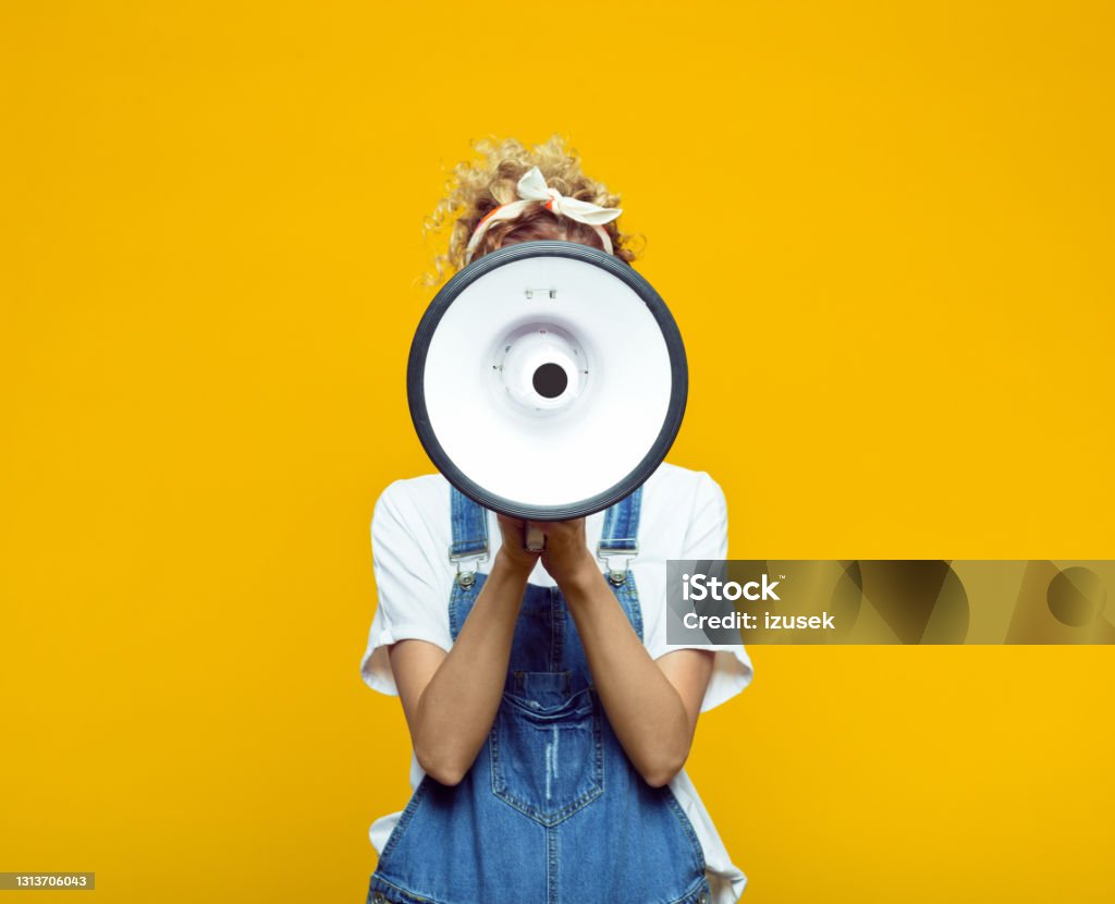 Young woman in dungarees shouting into megaphone Portrait of young woman wearing white t-shirt, denim dungarees and bandana shouting into megaphone. Studio shot on yellow background, unrecognizable person. Megaphone Stock Photo